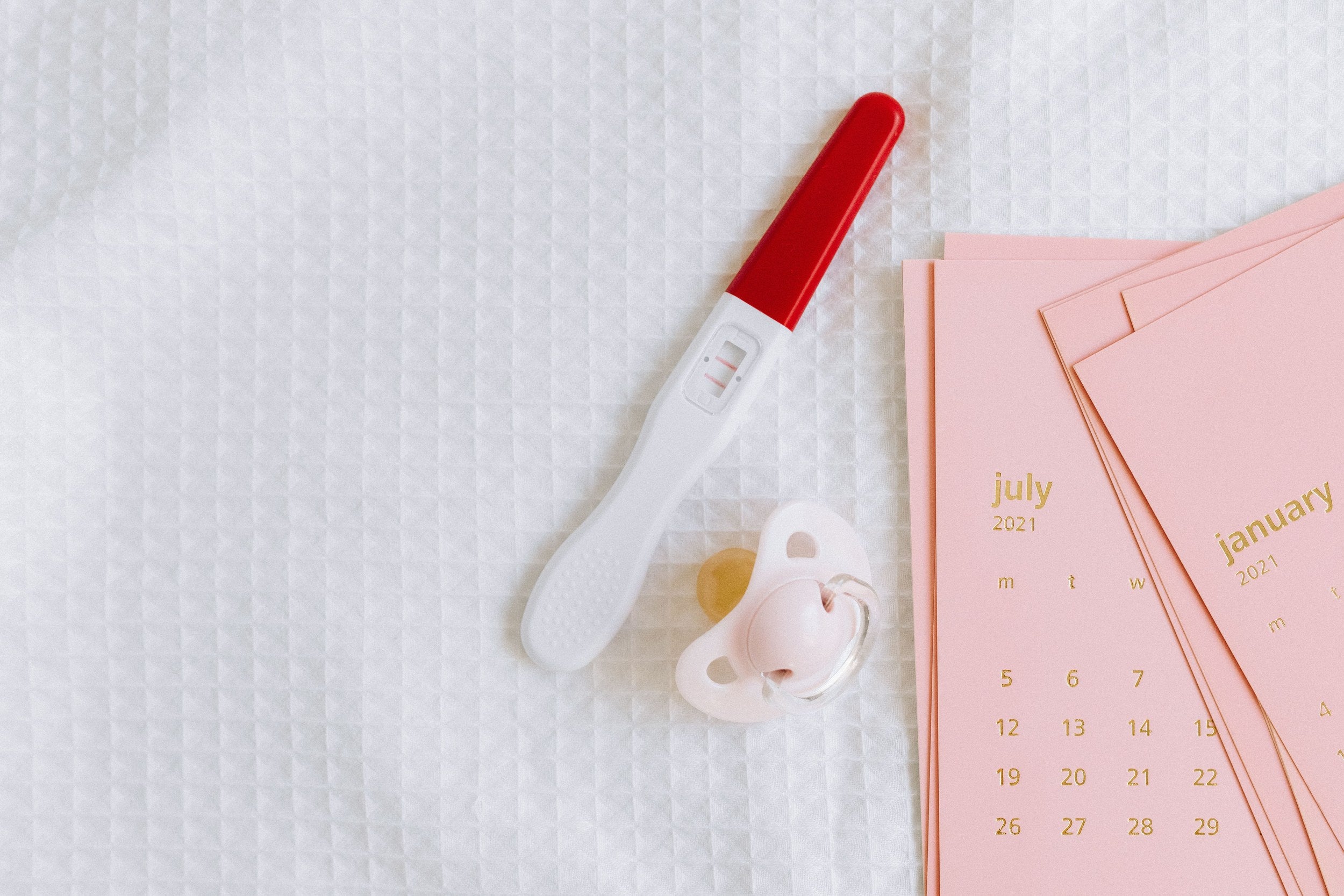 A pregnancy test, pacifier, and pink calendars on a white surface